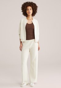 Kvinde med krøllet hår iført en cremefarvet skjortejakke med knapper, brun tanktop, cremefarvede bukser med vide ben, hvide sneakers og en guldkæde.