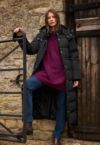 Kvinde i sort lang puffjakke, lilla sweater, blå jeans og brune støvler står på stentrappen og holder et sort metalhegn mod en stenvæg.