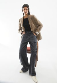 Hellbrauner strukturierter Blazer, schwarzes Oberteil und hoch taillierte dunkelgraue weite Jeans. Die Schuhe sind cremefarben und haben ein minimalistisches Design.