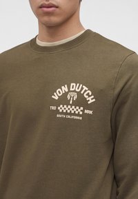 Gros plan d'une personne portant un sweat-shirt vert olive avec le logo beige « Von Dutch South California » et un motif à carreaux sur la poitrine.