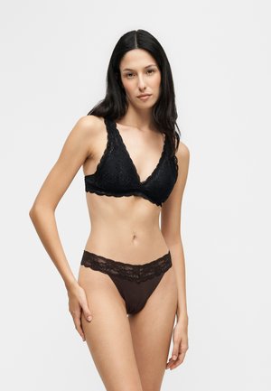 Donna con lunghi capelli scuri che indossa un bralette in pizzo nero e slip coordinati con rifiniture in pizzo nero, in piedi su uno sfondo chiaro e uniforme.