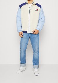 Giacca foderata in pile con corpo crema, maniche blu e colletto blu navy. Presenta una chiusura a pressione e dettagli del logo sul petto. Stile casual.
