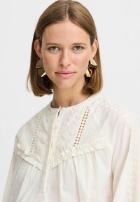 Weißes Blusenshirt mit Rüschendetails, texturierter Stickerei und Knopfleiste. Große goldene geometrische Ohrringe ergänzen den Look.