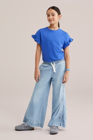 Tüdruk seisab, kandes sinist volangidega varrukatega särki, helehalli laia säärega pükse külgsete nööpidega, sinist kella ja mustrilisi libisevaid kingi.