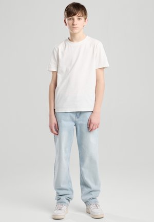 Tienerjongen staand tegen een grijze achtergrond, gekleed in een wit T-shirt, lichtblauwe spijkerbroek en witte sneakers, kijkt recht vooruit.