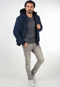 Marineblå dunjakke med hætte og lynlås foran, grå mønstret sweater indeni, lysegrå jeans og hvide sneakers.