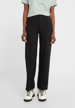 Personne portant un pantalon noir droit, une chemise à manches courtes vert clair et des bottes blanches à lacets, debout sur un fond gris clair.
