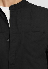 Chemise noire à manches longues avec col montant, motif texturé subtil, poche poitrine unique et boutons noirs.