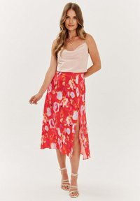 Světle růžový nátělník spojený s červenou sukní s květinovým vzorem a bočním rozparkem. Outfit doplňují béžové sandály na ramínka.
