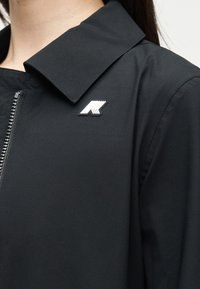 Κοντινό ενός ατόμου που φοράει μαύρο μπουφάν με διπλωμένο γιακά και ασημένιο καρφίτσα λογότυπου στην αριστερή πλευρά κοντά στο φερμουάρ.