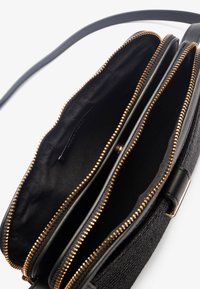 Schwarze Umhängetasche mit strukturiertem Äußeren, ausgestattet mit goldenen Reißverschlussverschlüssen und drei Fächern, die mit schwarzem Stoff ausgekleidet sind. Einfaches Gurt-Design.