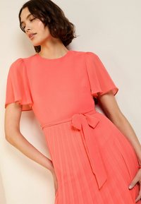 Korallenfarbenes plissiertes Kleid mit kurzen Puffärmelchen, einem runden Ausschnitt und einem Taillengürtel für eine optimale Passform. Glatte Textur mit fließender Silhouette.