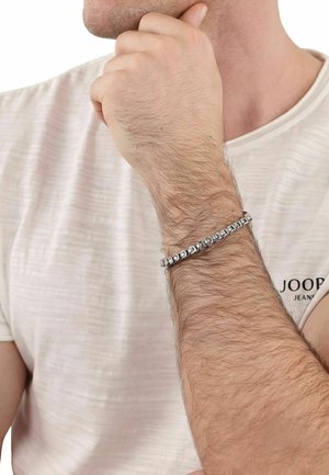 Uomo con maglietta bianca e braccio peloso, poggia il mento sulla mano chiusa a pugno, indossando un braccialetto a catena argentata al polso.