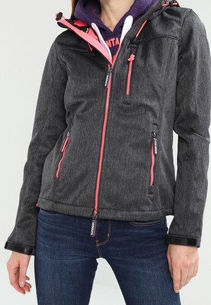 Kvinde iført en mørkegrå lynlåstrøje med pink lynlåse over en lilla hættetrøje og blå jeans, stående mod en ensfarvet baggrund.