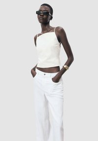 White sleeveless top with square neckline paired with white wide-leg pants. Model wears black sunglasses and a gold bangle. Smooth fabric texture.