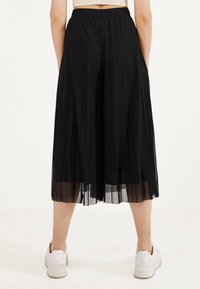 Zwarte geplooide midi-rok met een doorzichtige overlay, elastische tailleband en een zwierige silhouet, gecombineerd met witte sneakers.