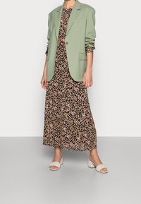 Lys grøn blazer med skulderpuder, parret med en sort blomstrede maxikjole med lyserøde og gule blomster, samt hvide blokhælede sandaler.