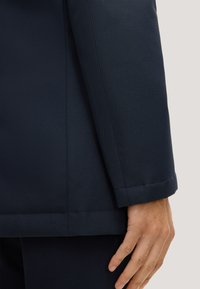 Donkerblauwe op maat gemaakte blazer met een gladde textuur, gestructureerde vorm en nauwkeurige stiksels; manchet zichtbaar, hand rustend op de stof.