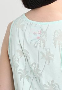Retro di una persona che indossa una maglietta senza maniche di colore azzurro chiaro con un motivo di palme grigie e una piccola toppa vicino al collo.