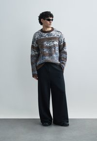 Stickad tröja med bruna, blå och vita mönster, rund halsringning och ribbade muddar, kombinerad med vida svarta byxor och solglasögon.