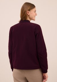 Woman smiling looking right, wearing a dark purple jacket and beige pants, shown from the back against a light beige background.