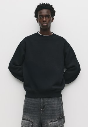 Zwarte sweatshirt met een ronde hals en lange mouwen, met een relaxte pasvorm. Draag met grijze shorts, tegen een effen witte achtergrond.