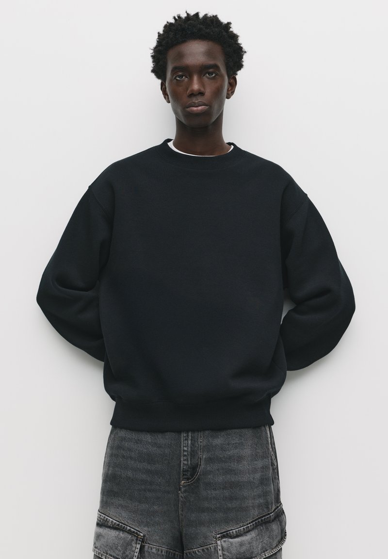 Schwarzer Sweater mit Rundhalsausschnitt und langen Ärmeln, mit einer lockeren Passform. Getragen mit grauen Shorts, vor einem schlichten weißen Hintergrund.