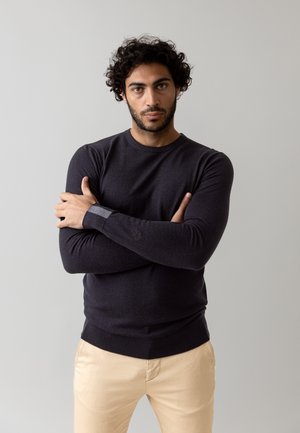 Pull en maille gris foncé à manches longues, col rond et poignets côtelés, associé à un chino beige clair. Texture lisse.