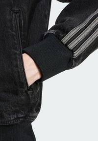 Svart denimjacka med detaljer som en ribbad svart manschett och ett tre-stripes silveraccent på axeln. Inkluderar en sidoficka.