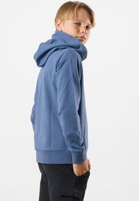 Felpa blu con tasca frontale, polsini a coste e cappuccio con cordino. Indossata con pantaloni cargo neri, il tessuto appare morbido e liscio.