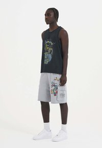 Black sleeveless tank top with a multicolored graphic, paired with light gray shorts featuring dragon designs and text accents. White sneakers.
