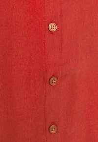 Camicia di lino rossa con una fila di quattro bottoni marroni, texture visibile e dettagli di cucitura sottili lungo la patta dei bottoni.