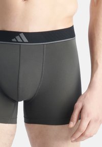 Herren Boxershorts aus dunkelgrünem Stoff. Sie verfügen über einen schwarzen Bund mit grauem adidas Logo und hellgrauen Nähten. Mittellange Ausführung.