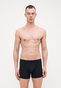 Sorte bomullsboxer med merket elastisk midje, tettsittende design og glatt tekstur. Modellen står foran en hvit bakgrunn.