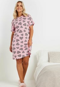 En rosa bomullsnattklänning med ett mönster av svarta hjärtan, korta ärmar och en sidoklyvning. Modellen bär mjuka rosa tofflor.