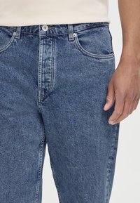 Main gauche à côté d'un jean en denim bleu avec bouton à l'avant, passants de ceinture et poches visibles, portée avec une chemise blanche rentrée.