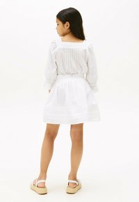Robe blanche à manches longues, corsage smocké, jupe évasée et accents brodés. Portée avec des sandales compensées blanches. Fond uni.