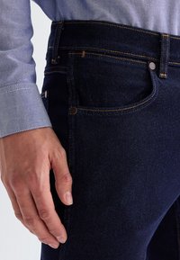 Artimas vaizdas žmogaus, dėvinčio tamsiai mėlynas džinsines kelnes ir šviesiai mėlyną ilgomis rankovėmis marškinius, matyti ranka, ilsėjusi šalia.