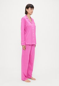 Ροδοκόκκινο σετ πιτζάμας με ρίγες που περιλαμβάνει ένα μπλουζάκι με κουμπιά και λευκές λεπτομέρειες, καθώς και παντελόνια με φαρδιά πόδια. Υφασμάτινη επιφάνεια με όρθιες ρίγες.