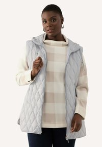 Ljusgrå, vadderad väst med dragkedja fram och huva, lager-på-lager över en krämfärgad och beige rutad topp. Slät textur, avslappnad design.