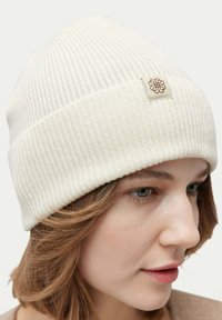 Gräddfärgad ribbad stickad mössa med en vikit bräm och en liten brun logotagg på sidan. Mjuk textur med en åtsittande passform.