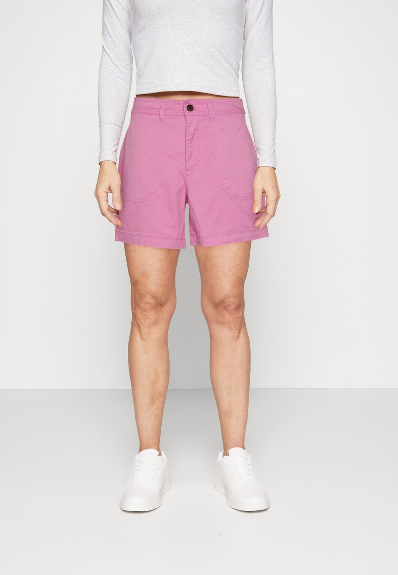 Shorts en coton rose avec une taille mi-haute, dotés de deux poches avant et d'une fermeture à bouton. Portés avec des baskets blanches et un haut à manches longues.