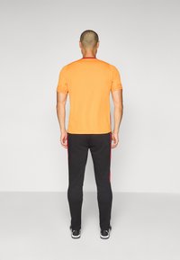 Orange atletisk t-shirt med mesh ærmer, parret med sorte jogger-bukser med røde sidelinder. Glat stof og tætsiddende design.