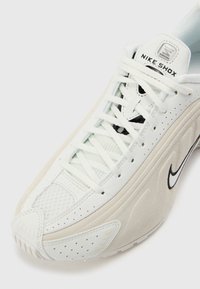Baskets blanches Nike Shox avec dessus en mesh et cuir, lacets blancs et swoosh noir Nike sur le côté.