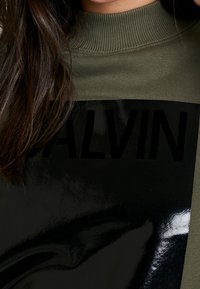 Olijfgroen trui met een glanzend zwart paneel waarop "CALVIN" in vet, zwarte letters over de borst is gedrukt en met geribbelde halsrand.
