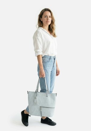 Frau steht, trägt weiße Bluse, hellblaue Jeans, schwarze Sneakers, hält große hellgraue Umhängetasche mit Quaste und vorderer Reißverschlusstasche.