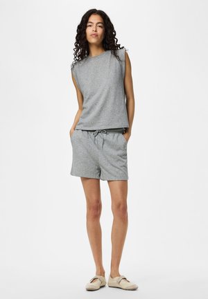 Femme aux cheveux bouclés portant un haut gris sans manches et un short assorti, debout les mains dans les poches devant un fond uni.