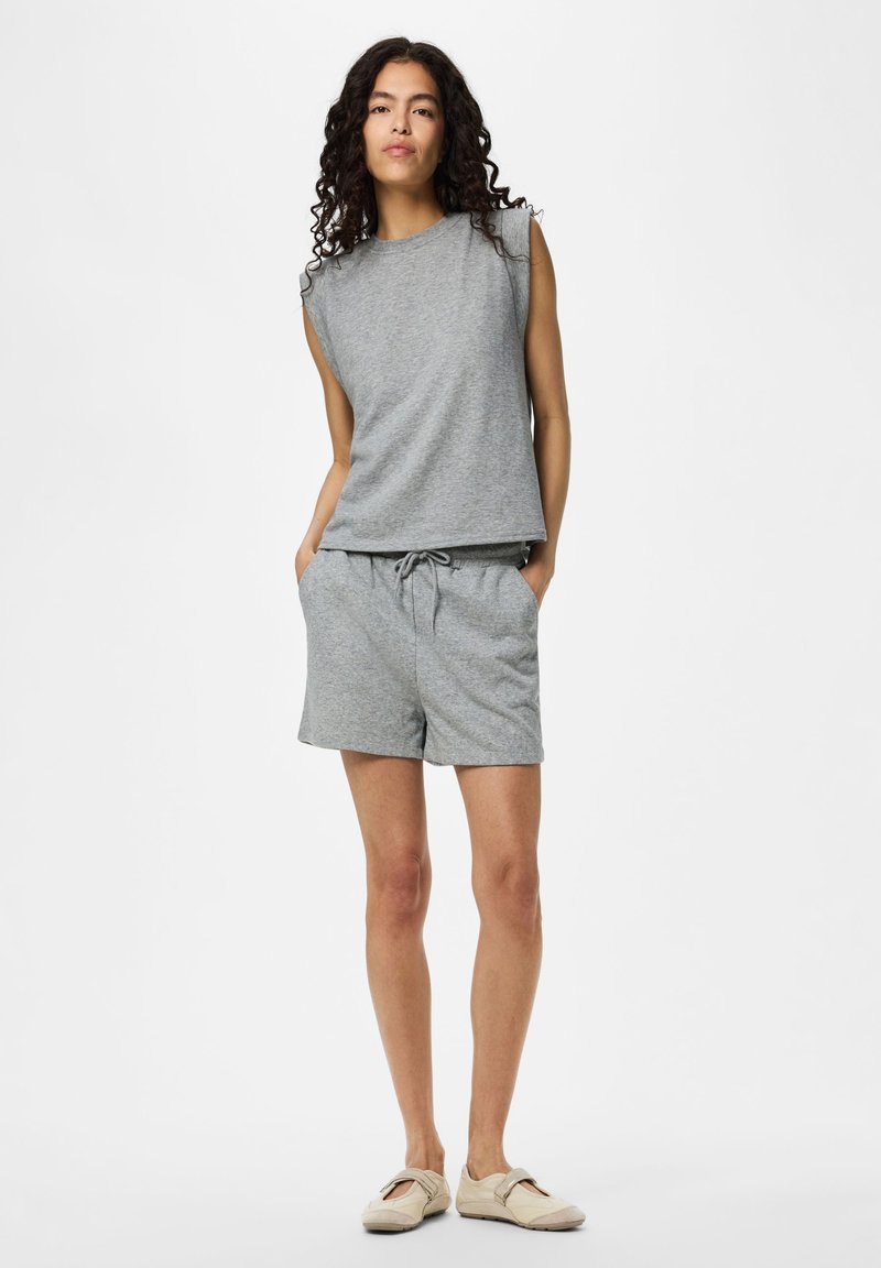 Femme aux cheveux bouclés portant un haut gris sans manches et un short assorti, debout les mains dans les poches devant un fond uni.
