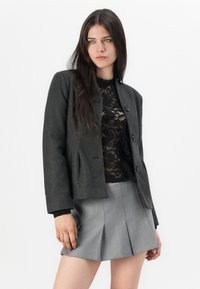 Chaqueta de mezcla de lana gris con un corte ajustado, cuello redondo y cuatro botones frontales, combinada con una blusa de encaje negra y una falda gris con plisados.