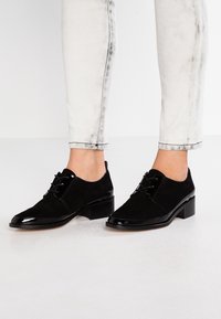 Czarne sznurowane buty wykonane z połączenia zamszu i polerowanego materiału lakierowanego, z lekko kwadratowym noskiem i niskim obcasem w kształcie bloku.